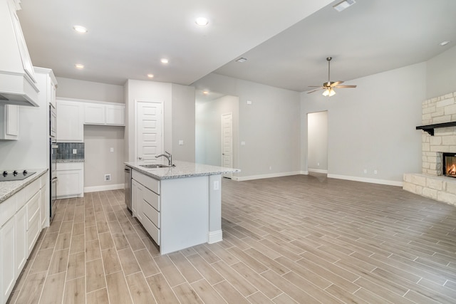 kitchen featuring tasteful backsplash, a stone fireplace, an island with sink, white cabinetry, and ceiling fan