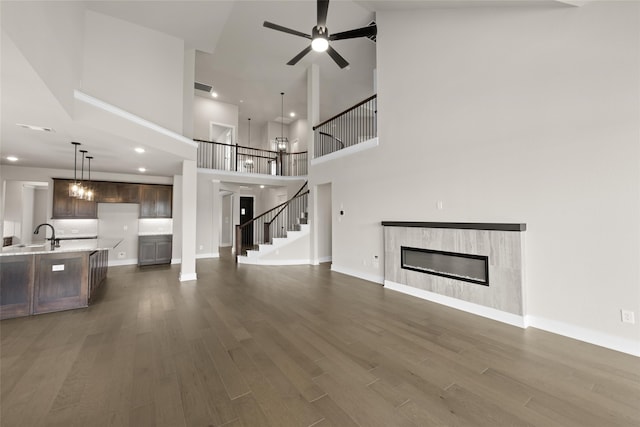 unfurnished living room with ceiling fan with notable chandelier, a tiled fireplace, dark wood-type flooring, sink, and a high ceiling