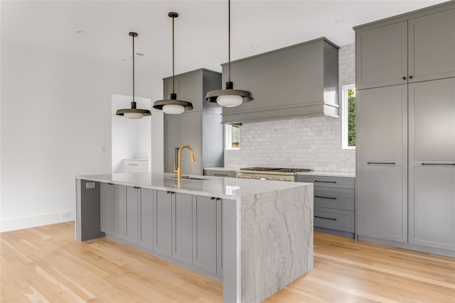 kitchen with light hardwood / wood-style flooring, tasteful backsplash, a center island with sink, premium range hood, and light stone countertops