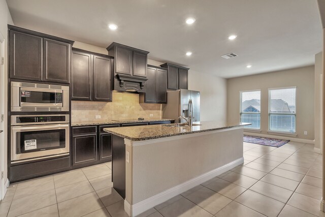 kitchen featuring tasteful backsplash, light stone counters, light tile patterned floors, a center island with sink, and stainless steel appliances