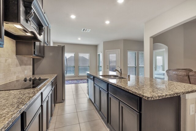 kitchen featuring black electric cooktop, custom exhaust hood, light stone counters, decorative backsplash, and an island with sink