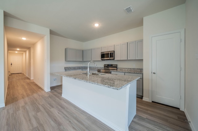 kitchen featuring gray cabinetry, a center island with sink, light wood-type flooring, stainless steel appliances, and light stone countertops