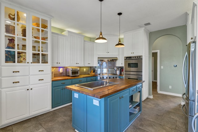 kitchen with wooden counters, blue cabinets, pendant lighting, a kitchen island with sink, and appliances with stainless steel finishes