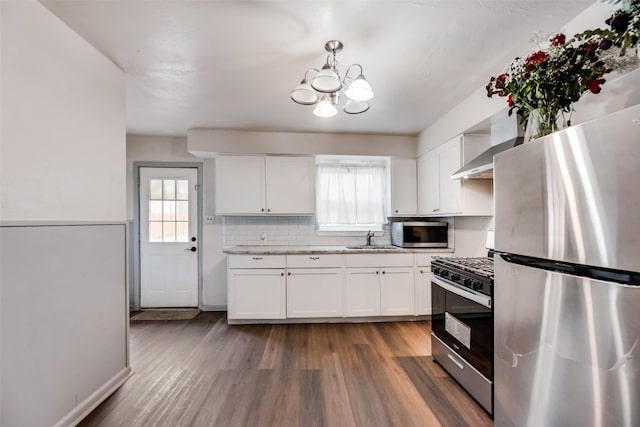 kitchen with appliances with stainless steel finishes, a chandelier, wall chimney exhaust hood, white cabinetry, and light wood-type flooring