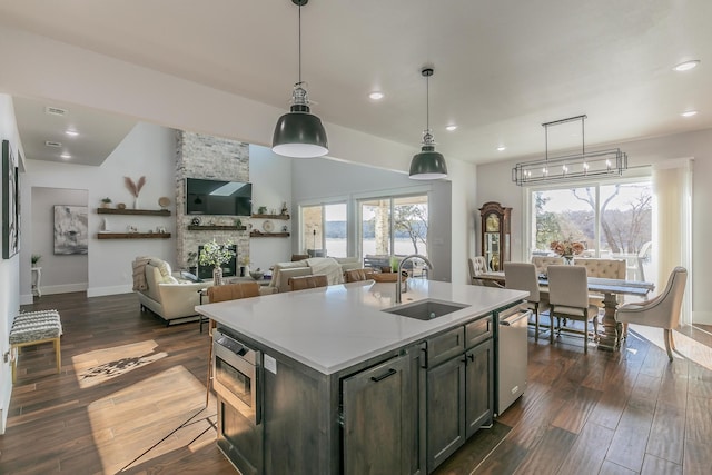 kitchen with a stone fireplace, a center island with sink, dishwasher, dark wood-type flooring, and sink