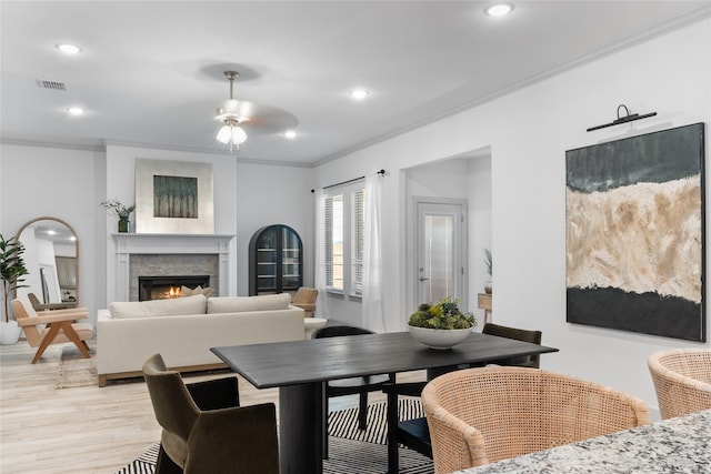 dining area featuring ceiling fan, a stone fireplace, light hardwood / wood-style flooring, and crown molding