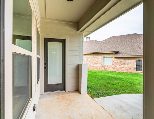 view of exterior entry with a yard, a shingled roof, and brick siding