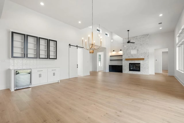 unfurnished living room with ceiling fan with notable chandelier, a large fireplace, light wood-type flooring, wine cooler, and a towering ceiling