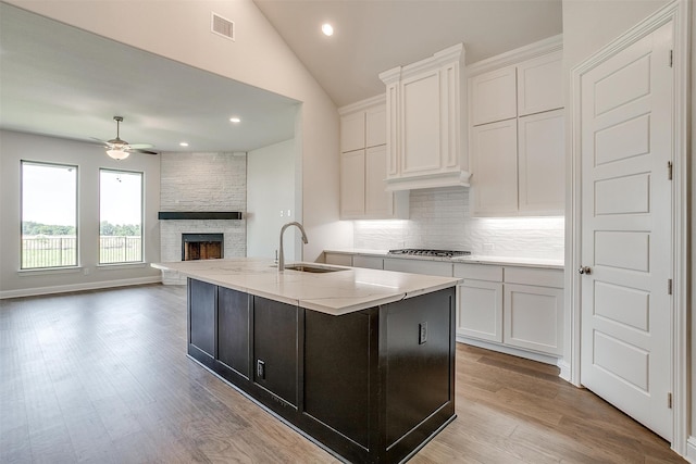 kitchen with white cabinetry, a stone fireplace, ceiling fan, a center island with sink, and sink