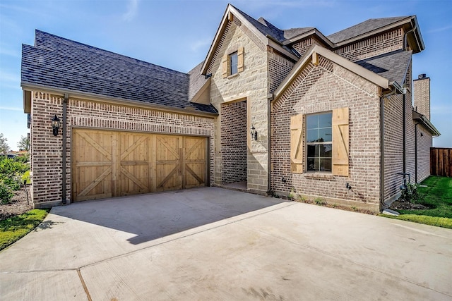 french country home featuring driveway, a garage, stone siding, a chimney, and brick siding