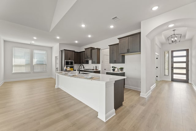 kitchen featuring stainless steel microwave, an inviting chandelier, light hardwood / wood-style flooring, a center island with sink, and dark brown cabinets