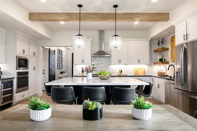 kitchen featuring a barn door, beamed ceiling, stainless steel appliances, wall chimney exhaust hood, and decorative light fixtures