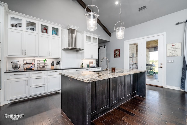 kitchen with a kitchen island with sink, dark wood-type flooring, hanging light fixtures, wall chimney exhaust hood, and dark stone countertops