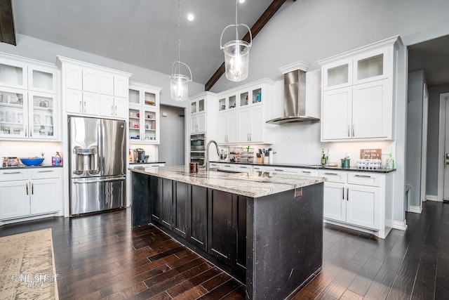 kitchen featuring wall chimney range hood, dark stone countertops, pendant lighting, a center island with sink, and appliances with stainless steel finishes