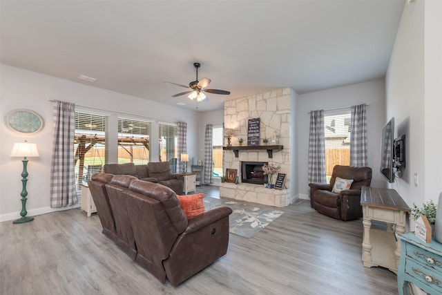 living area featuring a ceiling fan, a stone fireplace, light wood-style flooring, and baseboards