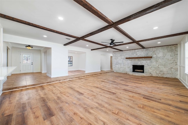 unfurnished living room featuring ceiling fan, light hardwood / wood-style floors, beamed ceiling, and a stone fireplace