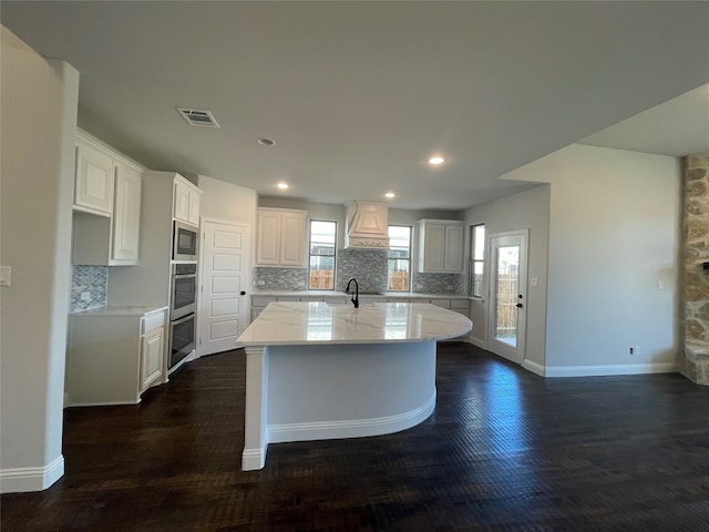 kitchen featuring built in microwave, white cabinetry, an island with sink, and custom range hood