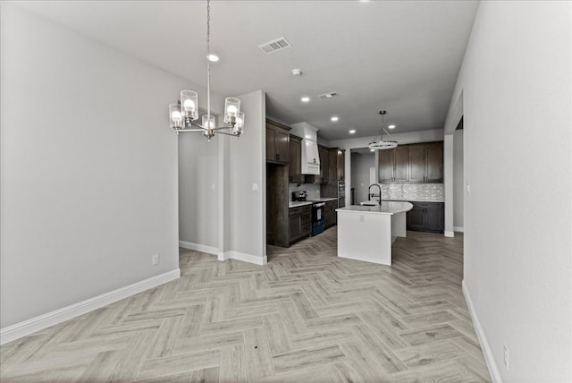 kitchen with light parquet floors, decorative light fixtures, backsplash, a center island with sink, and dark brown cabinetry