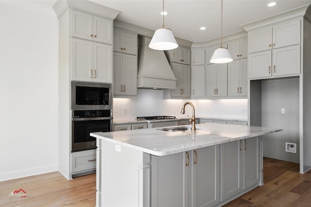 kitchen featuring an island with sink, premium range hood, stainless steel appliances, light stone countertops, and backsplash