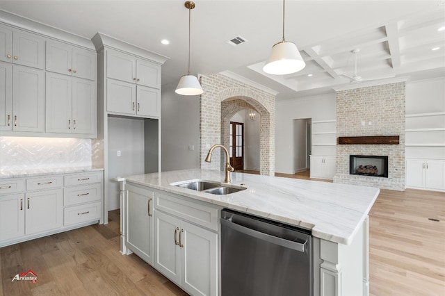 kitchen featuring coffered ceiling, light hardwood / wood-style floors, a brick fireplace, a center island with sink, and dishwasher