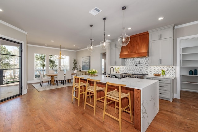 kitchen featuring light stone countertops, hardwood / wood-style floors, custom range hood, hanging light fixtures, and a kitchen island with sink