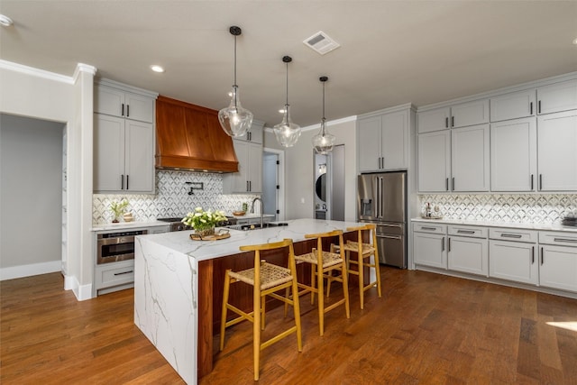 kitchen featuring light stone countertops, appliances with stainless steel finishes, an island with sink, wood-type flooring, and custom exhaust hood