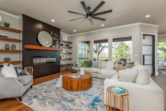 living room with wood-type flooring, a large fireplace, ceiling fan, and crown molding