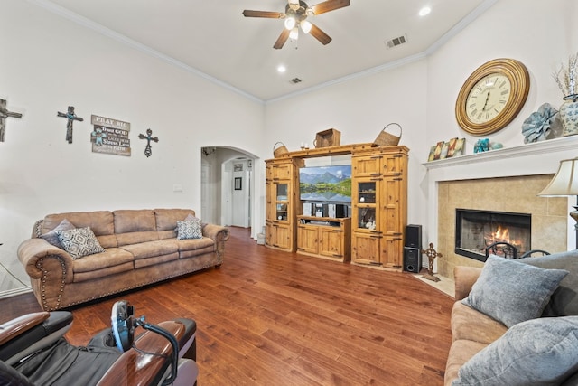 living room with a tiled fireplace, a towering ceiling, crown molding, wood-type flooring, and ceiling fan