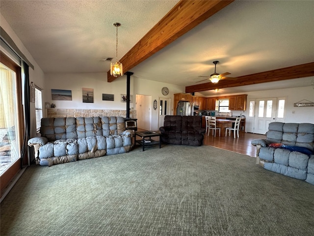 carpeted living room featuring ceiling fan, a wood stove, a textured ceiling, and beam ceiling