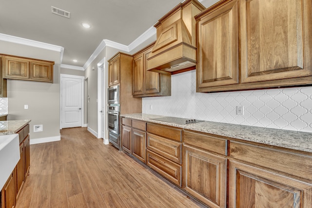 kitchen with light hardwood / wood-style flooring, custom range hood, crown molding, light stone countertops, and appliances with stainless steel finishes