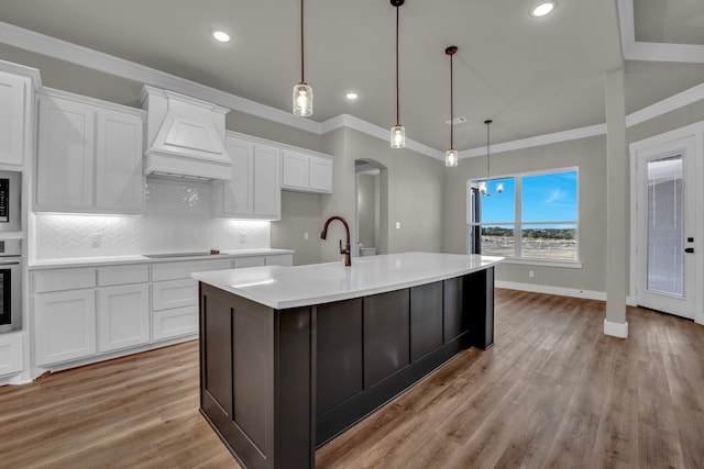 kitchen featuring premium range hood, a kitchen island with sink, decorative light fixtures, white cabinets, and light hardwood / wood-style flooring