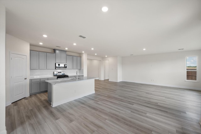 kitchen featuring light wood-type flooring, backsplash, stainless steel appliances, a center island with sink, and gray cabinets