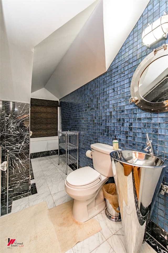 bathroom featuring toilet, marble finish floor, vaulted ceiling, and tile walls