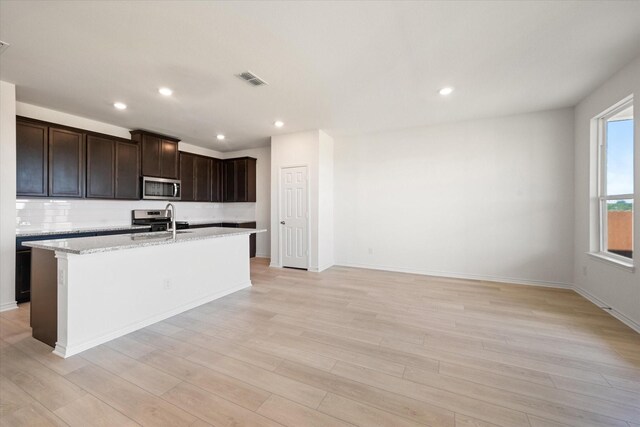 kitchen featuring tasteful backsplash, light hardwood / wood-style flooring, stainless steel appliances, light stone counters, and a center island with sink