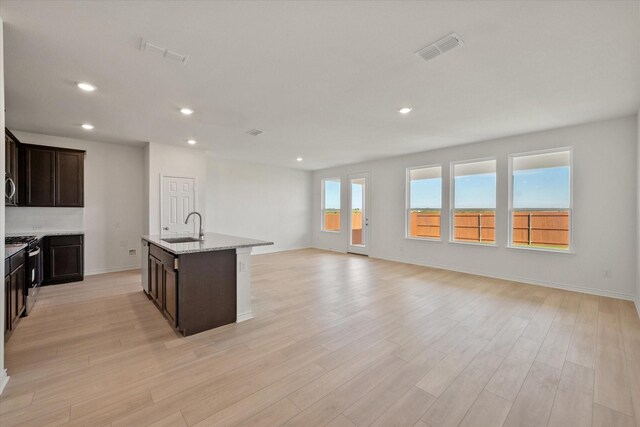 kitchen with stainless steel range with gas stovetop, light wood-type flooring, light stone counters, sink, and a center island with sink