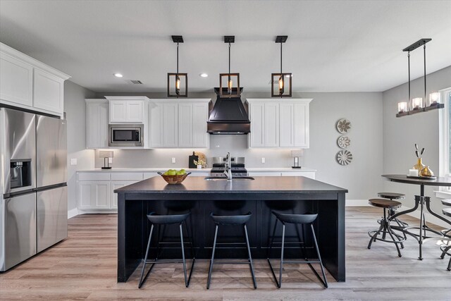 kitchen featuring white cabinetry, stainless steel appliances, premium range hood, a center island with sink, and light hardwood / wood-style floors