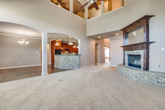 unfurnished living room featuring a towering ceiling, a brick fireplace, dark colored carpet, ceiling fan with notable chandelier, and ornamental molding