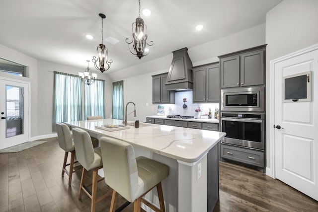 kitchen with an inviting chandelier, appliances with stainless steel finishes, premium range hood, a center island with sink, and dark hardwood / wood-style flooring