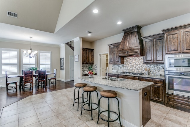 kitchen with a kitchen island with sink, stainless steel appliances, ornamental molding, sink, and a notable chandelier