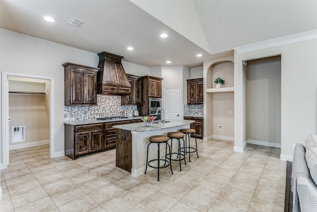 kitchen with an island with sink, appliances with stainless steel finishes, custom range hood, a breakfast bar, and decorative backsplash