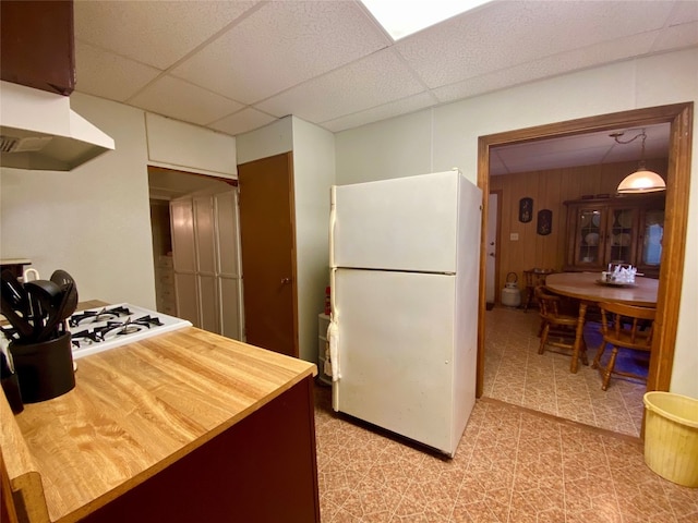 kitchen featuring decorative light fixtures, light tile flooring, a drop ceiling, white appliances, and wall chimney exhaust hood