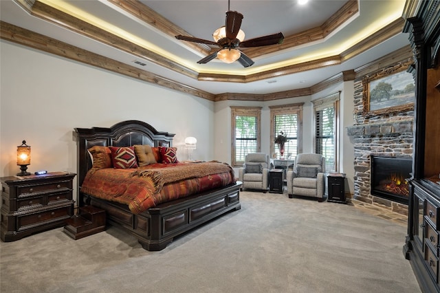 carpeted bedroom with ceiling fan, a stone fireplace, a raised ceiling, and ornamental molding