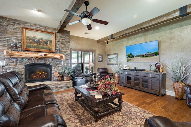 living room featuring ceiling fan, beamed ceiling, light hardwood / wood-style flooring, a textured ceiling, and a stone fireplace