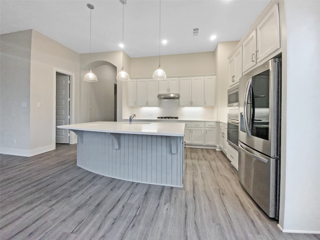 kitchen with an island with sink, pendant lighting, stainless steel appliances, white cabinets, and light wood-type flooring