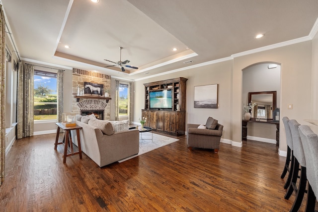 living room featuring dark hardwood / wood-style flooring, a raised ceiling, ceiling fan, crown molding, and a stone fireplace