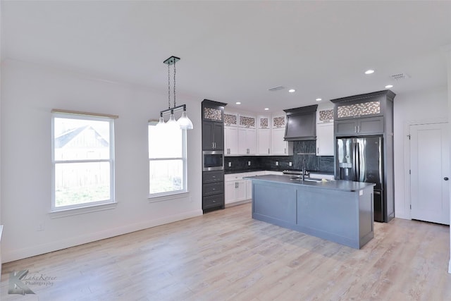 kitchen featuring light hardwood / wood-style floors, a kitchen island with sink, backsplash, premium range hood, and hanging light fixtures