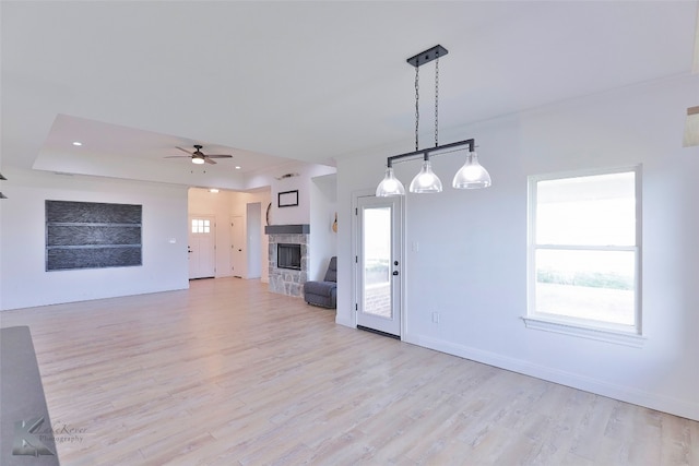 unfurnished living room featuring a raised ceiling, ceiling fan, a stone fireplace, and light hardwood / wood-style flooring