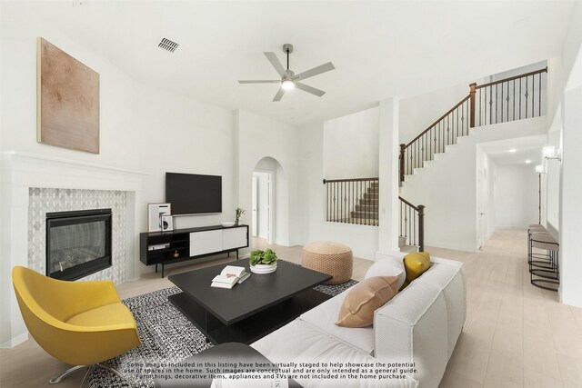 living room featuring a towering ceiling, ceiling fan, a tiled fireplace, and light hardwood / wood-style flooring