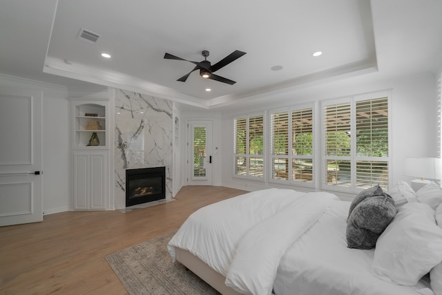 bedroom featuring light wood-type flooring, a raised ceiling, ceiling fan, crown molding, and a premium fireplace