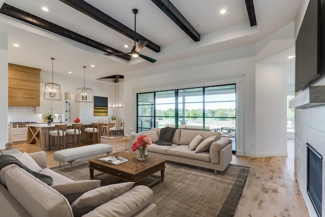 living room with a tile fireplace, ceiling fan with notable chandelier, beam ceiling, and light hardwood / wood-style flooring